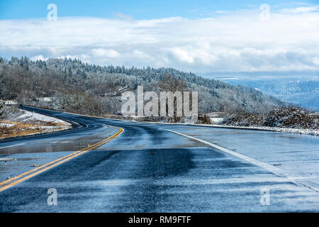 Paesaggio invernale con fusione umida su strada e colline coperte di neve alberi, prati e la strada alle spalle e montagne nascosti nelle nuvole appesi in Foto Stock