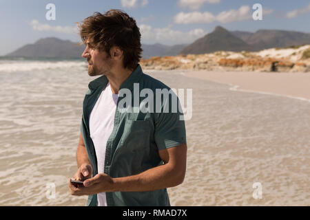 Giovane uomo che guarda lontano mentre si utilizza il telefono cellulare sulla spiaggia Foto Stock