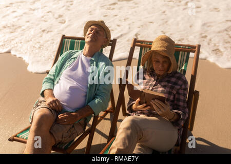 Senior donna leggere un libro mentre uomo senior relax sulle sdraio in spiaggia Foto Stock