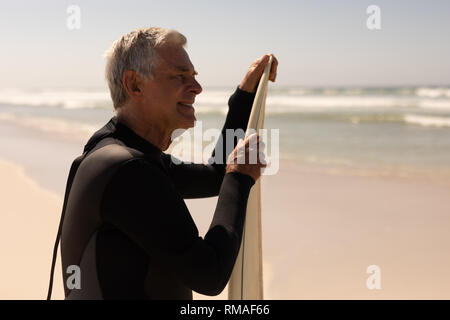 Vista laterale del senior surfista maschio in piedi con la tavola da surf in spiaggia Foto Stock