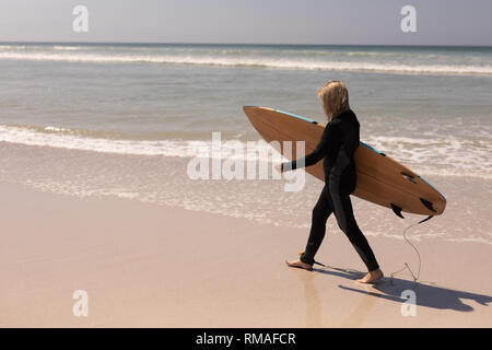 Vista laterale del senior surfista femmina a piedi con la tavola da surf in spiaggia Foto Stock