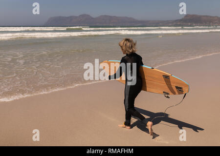 Vista laterale del senior surfista femmina a piedi con la tavola da surf in spiaggia Foto Stock