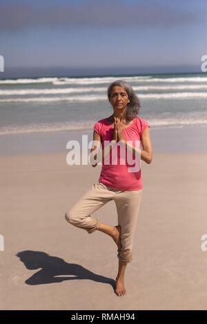 Senior donna con gli occhi chiusi e le mani incrociate fare yoga sulla spiaggia Foto Stock