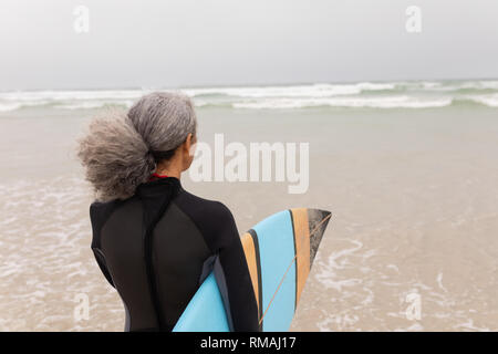 Senior surfista femmina a camminare verso il mare con la tavola da surf in spiaggia Foto Stock