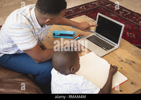Elevato angolo di vista americano africano padre aiutando il suo figlio con i compiti di un portatile a tavola Foto Stock