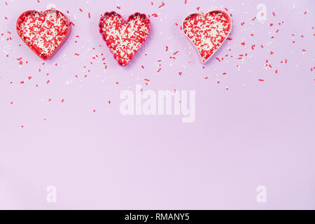 Una fila di a forma di cuore ad cookie cutters riempito con il rosso e bianco spruzza su un pallido sfondo viola Foto Stock