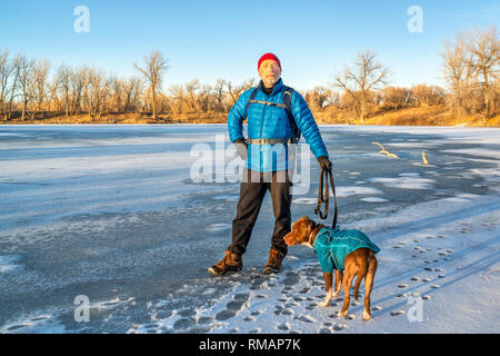 Senior escursionista maschio con un pitbull cane ama il tramonto sulla riva di un lago ghiacciato Foto Stock