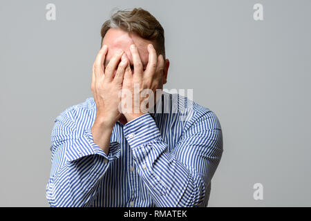 Uomo di mezza età che copre il viso con le mani in un corpo superiore ritratto su sfondo grigio Foto Stock