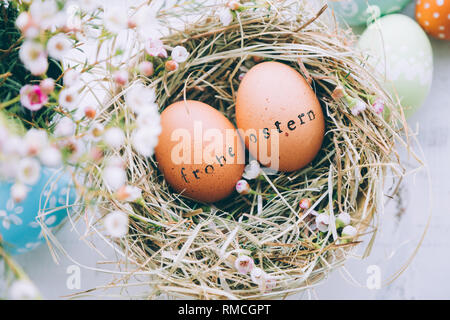 Le uova di pasqua con il timbro di gomma 'Frohe Ostern' Testo messaggio di saluto in un fieno e fiori su un bianco sullo sfondo di legno Foto Stock