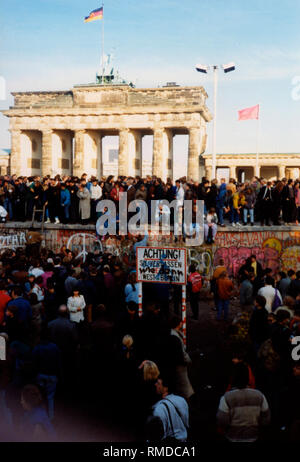 Una folla di persone che sorge di fronte e sul muro di Berlino. Sullo sfondo, la Porta di Brandeburgo. La foto è stata scattata poco dopo la caduta del muro di Berlino. Foto Stock