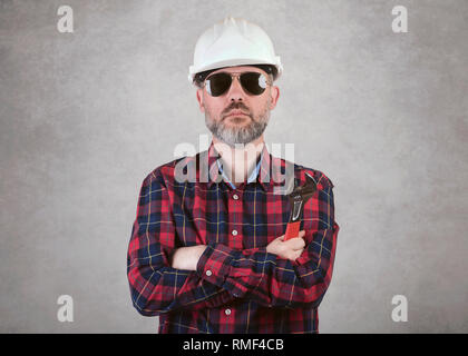 L'uomo lavoratore edile con un casco bianco e occhiali da sole tenere una chiave contro uno sfondo grigio Foto Stock