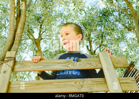 Bambino divertente sventolare sulla parte superiore della sua casa sulla struttura in legno su un albero di olivo, godendo della sua infanzia. Foto Stock