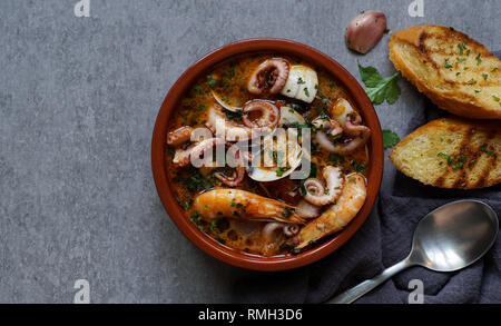 Da sopra una ciotola di zuppa di gamberetti, polpi e erbe vicino a cucchiaio, aglio e fette di pane su sfondo grigio Foto Stock