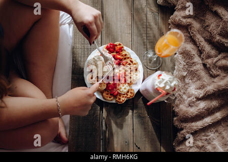 Giovane donna che mangia appetitosi waffle e gustose bevande sul vassoio a letto Foto Stock