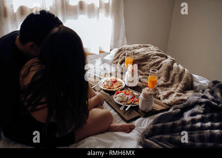 Vista posteriore del giovane avvolgente e seduta sul letto vicino al vassoio con deliziose cialde belghe con frutti di bosco e crema, bicchieri di succo di frutta fresco e frappè Foto Stock