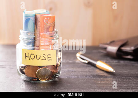 Un vaso di vetro con le monete e le fatture in euro con la parola casa su un tavoli in legno nero e sfondo. Penna e portafoglio in pelle. Il concetto di sav finanziario Foto Stock
