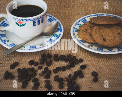 La prima colazione di una tazza in porcellana, un antico cucchiaio, biscotti al cioccolato e caffè in grani su una tela vintage background Foto Stock