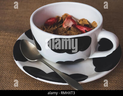 La prima colazione di un vaso di porcellana con cornflakes con bacche rosse e un cucchiaio di antiquariato su una tela vintage background Foto Stock