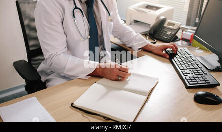 Le mani del medico maschio alla ricerca di nuove informazioni sui farmaci utilizzando computer desktop alla sua scrivania. Medical professional utilizzando il computer e scrivere note Foto Stock