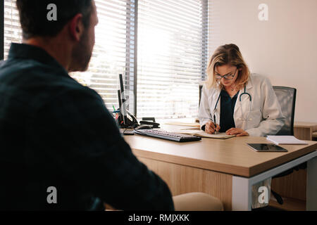 Medico donna rendendo note nel suo libro durante la consultazione di un paziente. Medico scrivendo la storia medica del paziente di sesso maschile seduto di fronte la sua scrivania in ospedale. Foto Stock