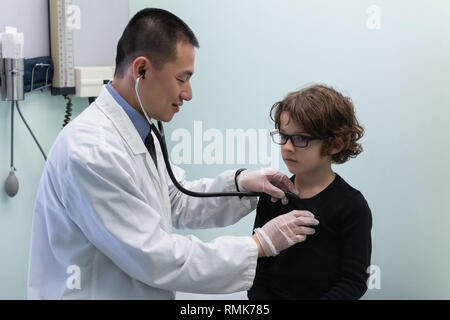 Giovani asiatici medico maschio esaminando ragazzo caucasico paziente con uno stetoscopio in clinica Foto Stock