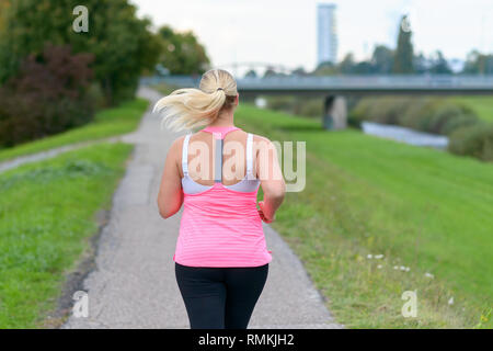 Vista posteriore della donna bionda di indossare abbigliamento sportivo che corre lungo il fiume in una vista ravvicinata Foto Stock