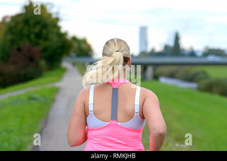 Vista posteriore della donna bionda di indossare abbigliamento sportivo che corre lungo il fiume in una vista ravvicinata Foto Stock