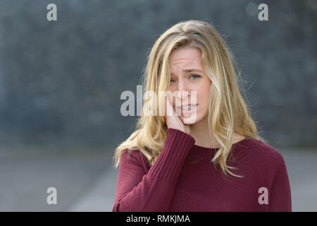 Triste giovane donna con un espressione malinconica in piedi tenendo la mano alla sua guancia come lei guarda la telecamera con spazio di copia Foto Stock