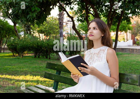 Bella scuola o college ragazza è seduta sul banco di lavoro con un libro in mano e pensando a qualcosa nel parco. Foto Stock