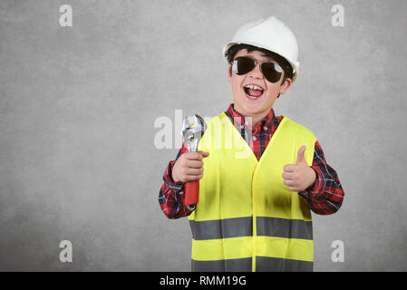 Bambino lavoratore edile nel casco bianco e occhiali da sole e tenere una chiave contro uno sfondo grigio Foto Stock