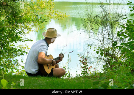 Giovane uomo a suonare la chitarra da un laghetto in estate Foto Stock