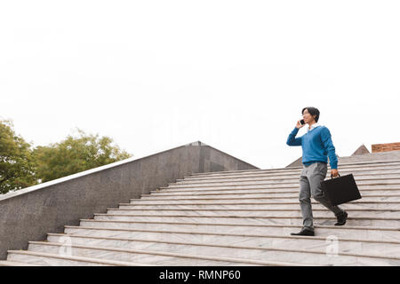 Sorridente giovane asian business uomo parlando al telefono cellulare mentre passeggiate all'aperto, portando nella valigetta Foto Stock