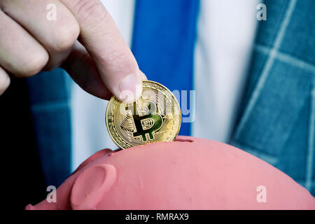 Primo piano di un giovane imprenditore caucasico, in un elegante abito grigio, inserendo un bitcoin in un maiale a forma di salvadanaio Foto Stock