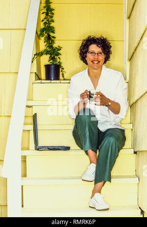 Un 30ish donna seduta sulla sua schiena passi da sorseggiando una tazza di caffè e di lavorare su un computer portatile. Foto Stock