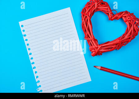 In vimini in legno cuore rosso e un vuoto di foglio bianco di notepad, accanto ad una matita, sfondo blu Foto Stock