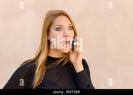 Donna attraente con un espressione sconvolta chat sul suo telefono cellulare con lo sguardo in avanti con la bocca agape e occhi grandi Foto Stock