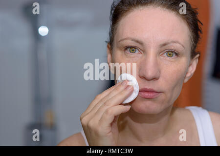 Donna che utilizza un tampone di cotone per pulire la sua pelle o rimuovere il trucco guardando la sua riflessione nello specchio di cura del viso e igiene personale concetto Foto Stock