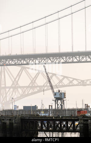 Il vecchio ponte di Forth Road con una gru in primo piano e il Ponte Forth Railway in background su un nebbioso giorno, Queensferry, Scozia Foto Stock