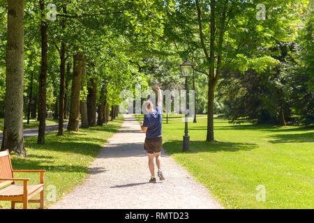 Uomo gioioso jogging in una boscosa verde parco di primavera in funzione lontano dalla telecamera punzonatura il pugno in aria come si gode il suo allenamento quotidiano Foto Stock