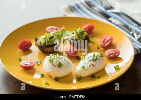 Uova al tegame con avocado farcito di funghi, pomodori e cipolle verdi su un allegro piatto giallo su un tavolo di legno Foto Stock