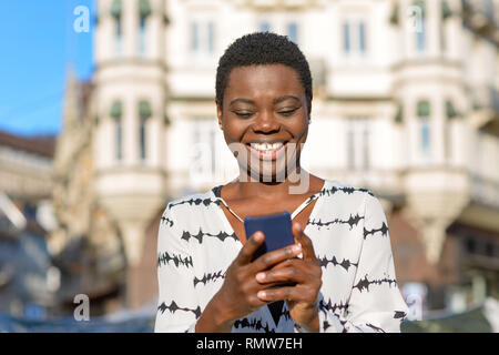 Felice elegante giovane donna africana ridere per il suo telefono cellulare come essa si erge in una strada di città in un basso angolo di visione contro un edificio storico Foto Stock