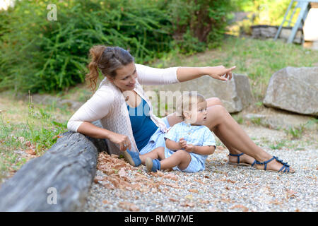 Giovane madre rilassante al di fuori con il suo bambino Figlio sulla terra che punta a qualcosa di fronte a lei con l'avviso toddler girando per visualizzarlo Foto Stock