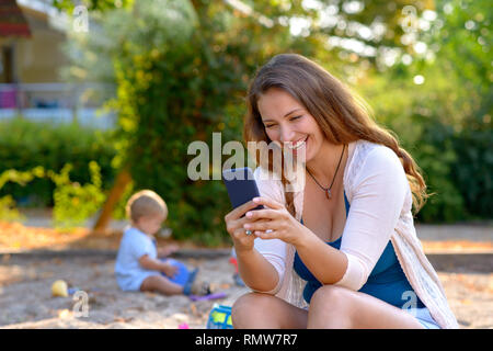 Vivace giovane madre ridere per un messaggio di testo sul suo telefono cellulare come lei siede all'aperto in un parco giochi osservando il suo Figlio bambino gioca con i suoi giocattoli Foto Stock