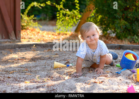 Avviso di carino piccolo bimbo gattona in sabbia in un parco giochi circondato dai suoi giocattoli di plastica come sorride felicemente con la videocamera Foto Stock