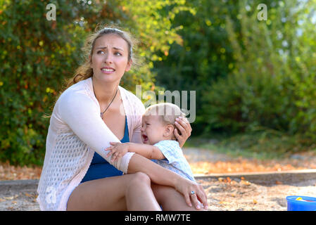 Sottolineato giovane madre la sua consolante Gridando bambino figlio guardando lontano con un angoscioso espressione come essi siedono insieme in un parco giochi all'aperto Foto Stock