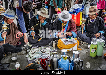 Tibetano etnica pregano gli uomini indossano abiti tradizionali durante la filatura palmare ruote della preghiera e di un soccorritore occasionale dà loro denaro. Foto Stock