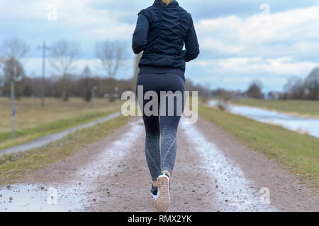 Montare sano giovane donna jogging lontano dalla telecamera in un angolo ritagliato vista su un paese bagnato via a fianco di un fiume in inverno Foto Stock