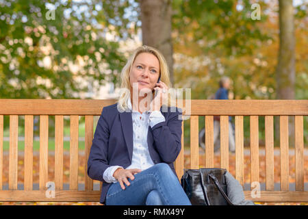 Attraente di mezza età donna bionda seduto su una panca di legno in una verdeggiante parco verde in chat sul suo cellulare Foto Stock