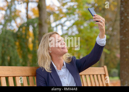 Attraente donna bionda con capelli tousled seduto su una tavola di legno panchina prendendo un selfie sul suo telefono cellulare Foto Stock