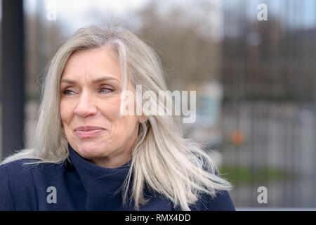 Attraente donna matura con spallamento tousled lunghezza capelli biondi guardando a sinistra del telaio con un lieto sorriso contro un sfocata backgrou urbana Foto Stock